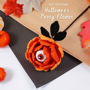 May include: A crocheted Halloween peony flower with an orange and white eye center. The flower is orange with black leaves and stem. The image also includes small pumpkins and fall leaves. Text on the image reads "PDF Pattern Halloween Peony Flower."