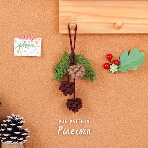 Könnte beinhalten: Handgefertigter Tannenzapfen-Schmuck mit grünen Blättern und drei braunen Tannenzapfen, die an einer braunen Schnur hängen. Der Hintergrund ist eine Pinnwand mit einer Holzleiste und dekorativen Elementen.