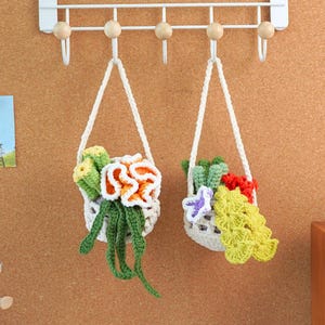 Puede incluir: Dos maceteros colgantes de crochet con flores de colores y vegetación. Los maceteros están hechos de hilo blanco y cuelgan de un estante de metal blanco.