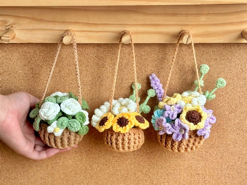 Op de afbeelding: Drie gebreide hangende bloempotten met verschillende bloemstukken. De potten zijn bruin en de bloemen zijn wit, geel en paars.