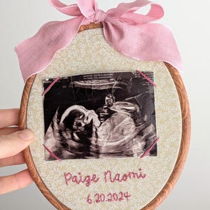 May include: Oval wooden embroidery hoop with a cream floral fabric background. A black and white ultrasound image is secured with pink thread. The text "Paige Naomi" and "6.20.2024" are embroidered in pink. A pink ribbon bow is at the top.