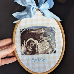 May include: An oval embroidery hoop with a baby ultrasound photo, light blue bow, and embroidered name "Lesty". The fabric is a beige and white gingham pattern. The wooden hoop is held by a hand.