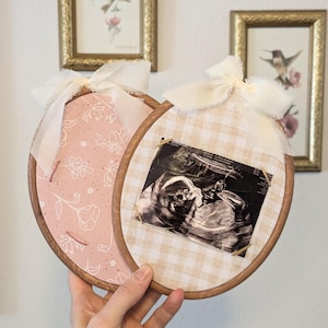 May include: Two oval wooden embroidery hoops used as frames. One hoop features pink fabric with a white floral line-art pattern. The other hoop contains beige gingham fabric holding a black and white ultrasound scan. Both hoops are topped with white ribbon bows.