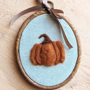 May include: An oval embroidery hoop with a wood-grain finish containing light blue fabric. A needle-felted orange pumpkin with a dark brown stem is centered on the fabric. A taupe-colored ribbon is tied in a bow at the top of the hoop.