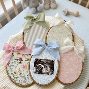 May include: Five oval wooden embroidery hoops containing fabric inserts with various patterns, including floral prints, stripes, and solid colors. Each hoop is topped with a decorative fabric bow. One hoop features a black and white ultrasound image.