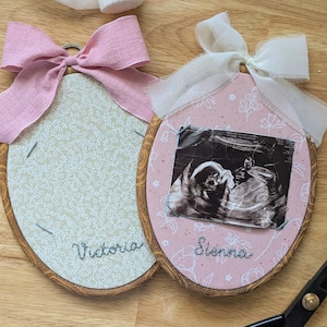 May include: Two oval wooden embroidery hoops featuring fabric inserts. One hoop has light green floral fabric with the name Victoria embroidered in gray. The other has pink floral fabric, a black and white ultrasound image, and the name Sienna embroidered in gray. Both hoops are topped with fabric bows.