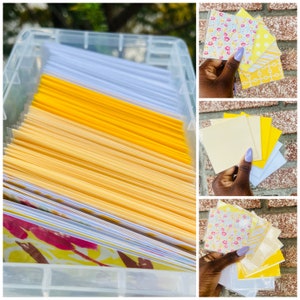 May include: A clear plastic storage container filled with stacks of yellow, white, and patterned paper cards. The cards are arranged in rows and columns. The container is sitting on a table.