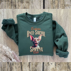 May include: A dark green sweatshirt with a graphic of a white sphinx cat with orange eyes. The text "Exclusive Anti-Social Sphinx Club" is printed in orange and white.