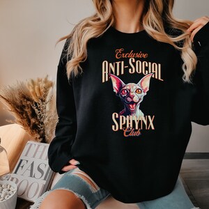 May include: Black sweatshirt with a graphic print of a white cat with orange eyes and the text "Exclusive Anti-Social Sphinx Club".