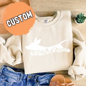 K&ouml;nnte beinhalten: Cremefarbenes Sweatshirt mit einer wei&szlig;en Dackel-Silhouette, gef&uuml;llt mit floralen Mustern. Das Wort "CUSTOM" ist auf einem orangefarbenen Kreis aufgedruckt. Das Sweatshirt wird mit blauen Jeans und Sandalen gezeigt.