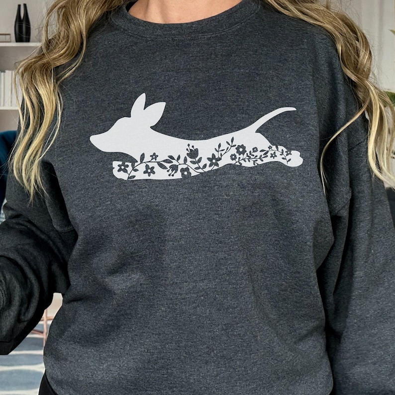 K&ouml;nnte beinhalten: Dunkelgraues Sweatshirt mit einer wei&szlig;en Hundesilhouette und floralen Details. Der Hund wird in Bewegung dargestellt, mit Blumen, die in seine Form integriert sind. Das Sweatshirt ist ein l&auml;ssiger, bequemer Stil.