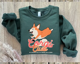 May include: A dark green sweatshirt with a graphic of a running corgi dog in orange and white. The text "Corgi Club 1934" is below the dog, with the phrase "In a world full of trends, be a corgi" underneath.
