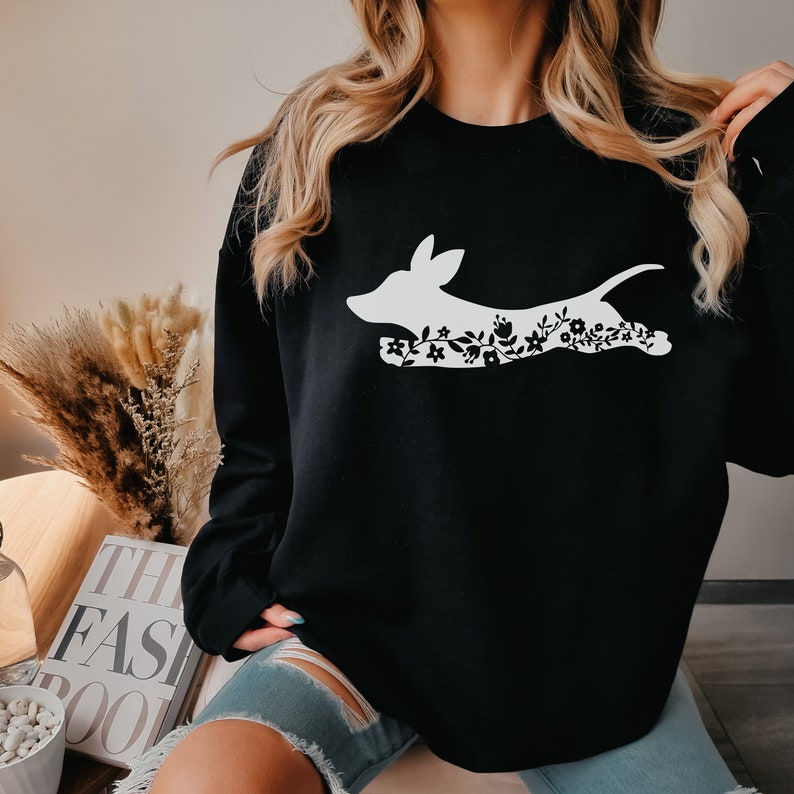 K&ouml;nnte beinhalten: Schwarzes Sweatshirt mit einer wei&szlig;en Silhouette eines Hundes, gef&uuml;llt mit floralen Mustern. Der Hund wird in einer Laufhaltung dargestellt. Das Sweatshirt hat eine einfarbige schwarze Farbe.