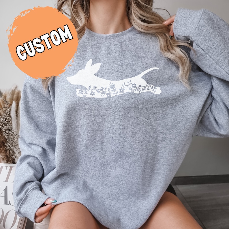 K&ouml;nnte beinhalten: Hellgraues Sweatshirt mit einem wei&szlig;en Hunde-Grafikmuster mit floralen Details. Das Wort "CUSTOM" ist wei&szlig; auf einem orangefarbenen Kreis gedruckt. Das Sweatshirt hat lange &Auml;rmel.