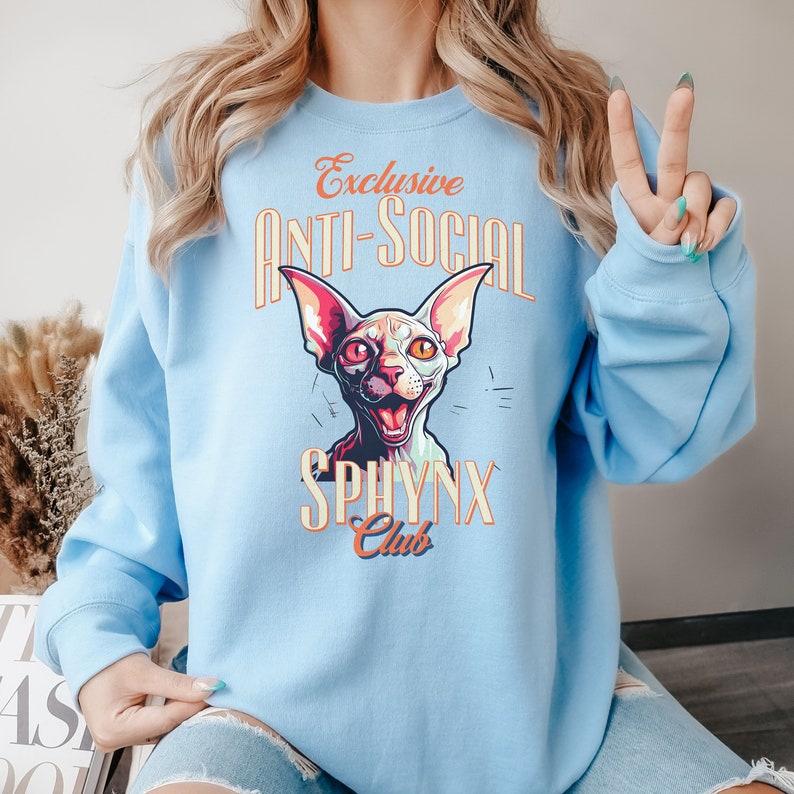 May include: Light blue sweatshirt with a graphic of a Sphynx cat with the text "Exclusive Anti-Social Sphynx Club" in orange and yellow.