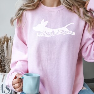 K&ouml;nnte beinhalten: Hellrosa Sweatshirt mit einem wei&szlig;en Aufdruck eines hunde&auml;hnlichen Wesens mit floralen Details. Das Sweatshirt hat lange &Auml;rmel und einen Rundhalsausschnitt. Das Design ist schlicht und feminin, geeignet f&uuml;r Freizeitkleidung.