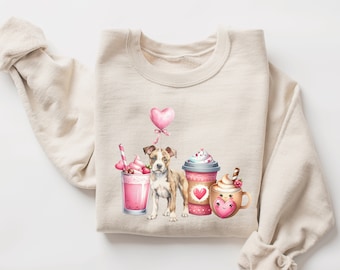 Puede incluir: Sudadera crema con una ilustración en acuarela de un perro, un globo de corazón rosa, un batido rosa y dos tazas de café. Las tazas y el batido tienen detalles de corazón y crema batida. Diseño temático de San Valentín.