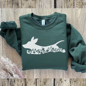 K&ouml;nnte beinhalten: Ein dunkelgr&uuml;nes Sweatshirt mit einem wei&szlig;en floralen Hunde-Design. Der Hund ist in einer Laufpose dargestellt, wobei das florale Design seinen K&ouml;rper ausf&uuml;llt. Das Sweatshirt hat einen Rundhalsausschnitt und lange &Auml;rmel mit umgeschlagenen Manschetten. Der Hintergrund ist eine hellbraune Holzoberfl&auml;che.
