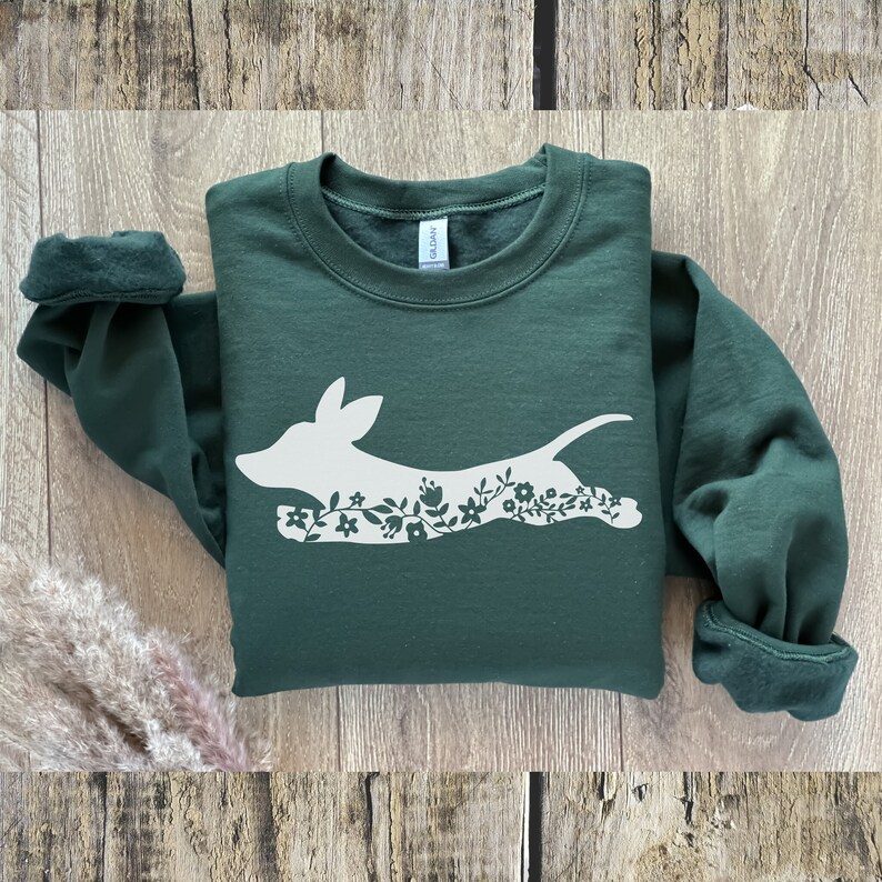 K&ouml;nnte beinhalten: Ein dunkelgr&uuml;ner Pullover mit einer wei&szlig;en Silhouette eines Dackels, der mit floralen Details rennt.