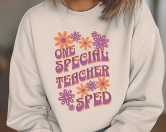Könnte beinhalten: Ein hellbeiger Pullover mit einem lila und orangefarbenen Grafikdesign. Die Grafik zeigt den Text "One Special Teacher SPED" in einer Retro-Schriftart mit Blumen, die den Text umgeben.