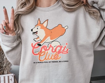 May include: A light gray sweatshirt with a graphic of a cartoon corgi dog jumping in the air. The graphic includes the text "Corgi Club" with the year "1934" in a circle. The text "In a world full of trends, be a corgi" is printed below the graphic.