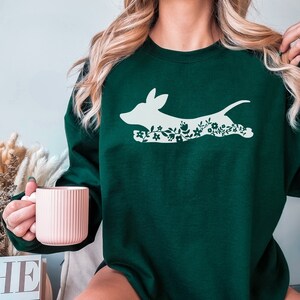K&ouml;nnte beinhalten: Ein dunkelgr&uuml;nes Sweatshirt mit einer wei&szlig;en Silhouette eines Hundes, verziert mit floralen Mustern. Der Hund ist in Bewegung dargestellt, mit Blumen und Bl&auml;ttern, die seinen K&ouml;rper bilden. Das Sweatshirt hat einen einfarbigen Hintergrund, und das Design ist mittig platziert.