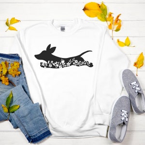 K&ouml;nnte beinhalten: Wei&szlig;es Sweatshirt mit einer schwarzen Silhouette eines Dackelhundes, verziert mit floralen Mustern. Der Hund wird in Bewegung dargestellt. Das Bild enth&auml;lt blaue Jeans und graue Turnschuhe.