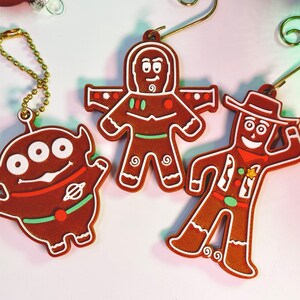 May include: Three gingerbread ornaments featuring characters from the Toy Story franchise. The ornaments are brown with white icing details and colourful accents. The characters are Buzz Lightyear, Woody, and Alien.