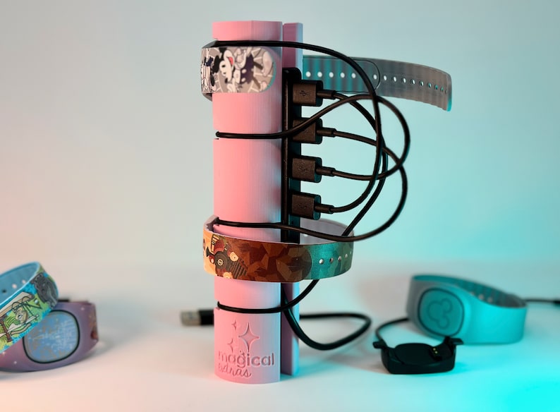 Pode incluir: Um suporte de carregamento rosa com o texto "magical extras" impresso no lado. O suporte tem quatro portas USB e est&aacute; segurando v&aacute;rias pulseiras coloridas com diferentes designs.