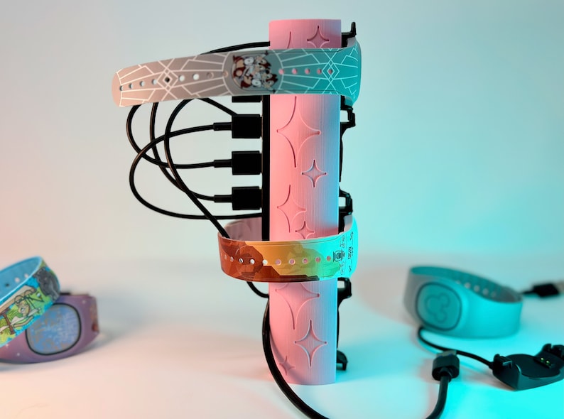 Pode incluir: Uma esta&ccedil;&atilde;o de carregamento rosa com quatro portas de carregamento e v&aacute;rios MagicBands Disney coloridos conectados a ela. Os MagicBands t&ecirc;m designs diferentes, incluindo um padr&atilde;o geom&eacute;trico, um design abstrato colorido e um design com um personagem Disney.