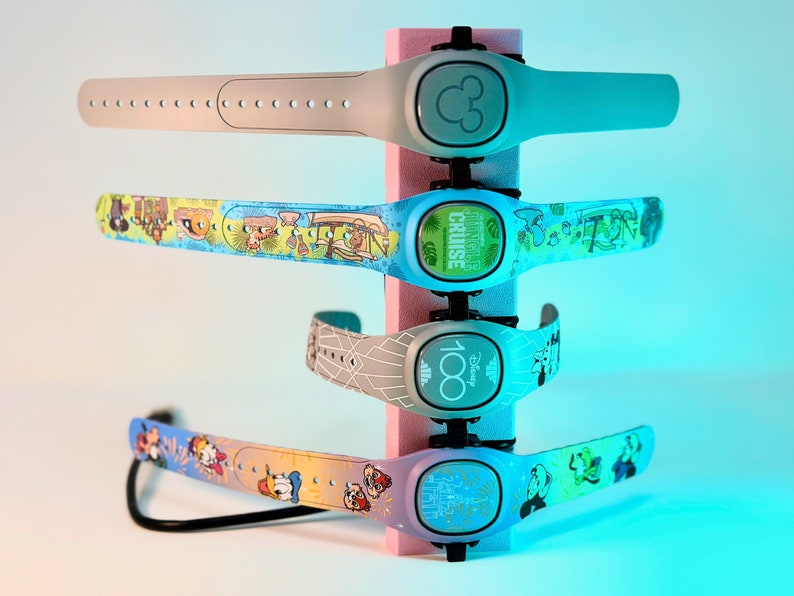 Pode incluir: Cinco MagicBands Disney coloridas com designs diferentes. A banda superior &eacute; cinza com um &iacute;cone do Mickey Mouse. A segunda banda &eacute; azul com um design de cruzeiro na selva. A terceira banda &eacute; cinza com um padr&atilde;o geom&eacute;trico branco e o texto "Disney 100". A quarta banda &eacute; amarela com um design do Mickey Mouse e seus amigos. A banda inferior &eacute; azul com um design de castelo de Cinderela.