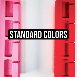 Pode incluir: Dois objetos impressos em 3D, um rosa fosco e outro vermelho fosco, com o texto "STANDARD COLORS" em letras pretas.