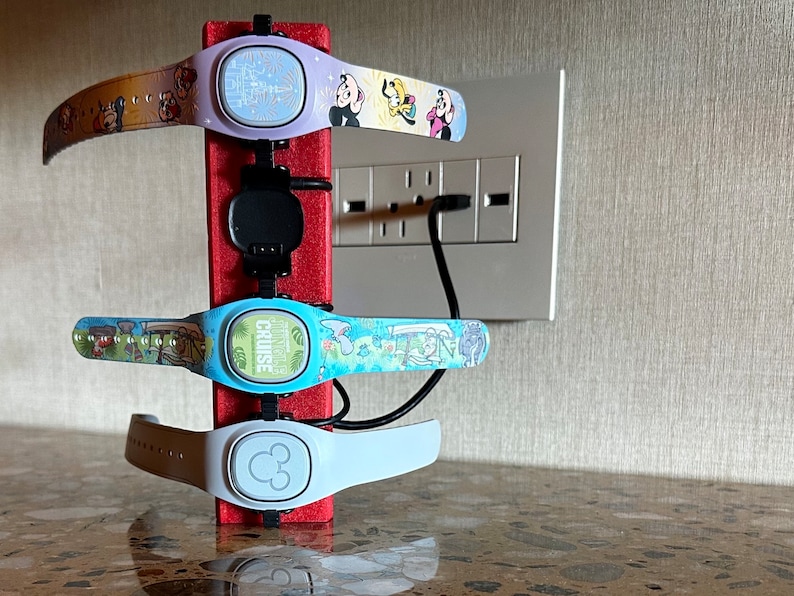 Pode incluir: Quatro MagicBands Disney carregando em um suporte de carregamento vermelho. As MagicBands s&atilde;o roxas, azuis, brancas e marrons com v&aacute;rios personagens e designs da Disney. O suporte de carregamento est&aacute; ligado a uma tomada de parede.