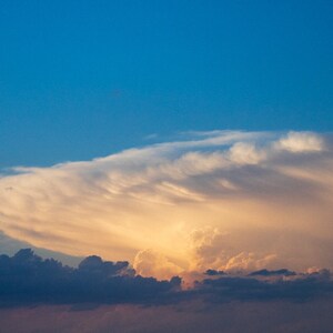 Puede incluir: Una vista panorámica de un cielo dramático con una gran nube esponjosa iluminada por el sol poniente. La nube es de un amarillo dorado suave con un toque de rosa, mientras que el resto del cielo es de un azul profundo.