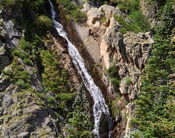 May include: A cascading waterfall flows down a rocky cliffside, surrounded by lush green trees and foliage. The water is clear and white, creating a beautiful contrast against the dark rocks.