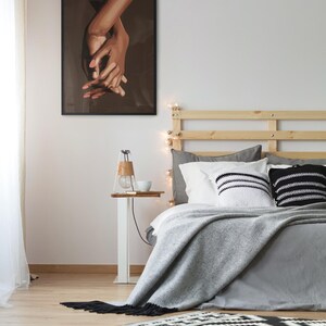 Puede incluir: Un dormitorio con una estructura de cama de madera, una manta gris y almohadas con estampado en blanco y negro. Una impresi&oacute;n enmarcada en negro de dos manos que se sostienen una a la otra cuelga de la pared sobre una mesita de noche blanca con una l&aacute;mpara y una taza.