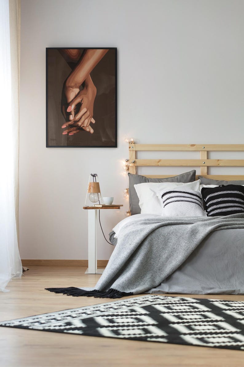 Puede incluir: Un dormitorio con una estructura de cama de madera, una manta gris y almohadas con estampado en blanco y negro. Una impresi&oacute;n enmarcada en negro de dos manos que se sostienen una a la otra cuelga de la pared sobre una mesita de noche blanca con una l&aacute;mpara y una taza.