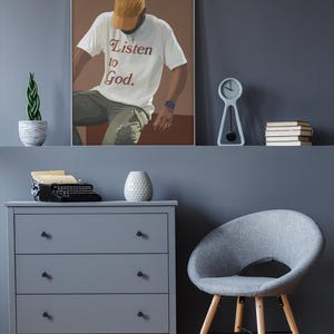 May include: A framed illustration of a person wearing a white t-shirt with the text "Listen to God." The person is wearing a yellow baseball cap and is sitting on a brown surface. The background is a light brown color.
