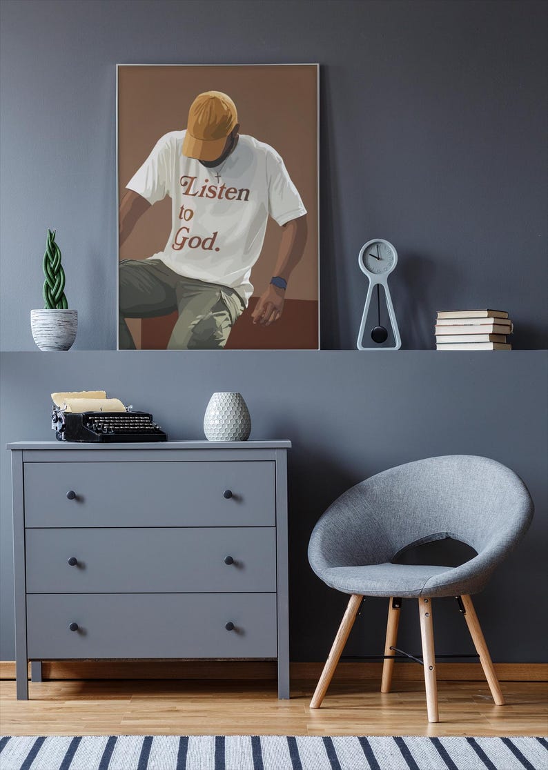 May include: A framed illustration of a person wearing a white t-shirt with the text "Listen to God." The person is wearing a yellow baseball cap and is sitting on a brown surface. The background is a light brown color.