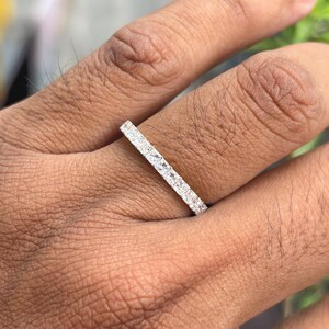May include: A silver-toned eternity band ring featuring a continuous row of small, square-cut clear stones set in a metallic channel. The ring is worn on a finger, showcasing the uniform alignment of the stones along the entire visible band.