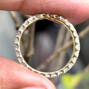 May include: A yellow gold eternity band featuring a continuous row of small, clear, round-cut gemstones set in a shared-prong setting. The ring is circular, showcasing a polished metallic finish and uniform stone placement around the entire band.