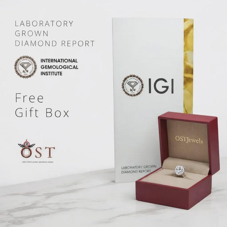 May include: A round-cut diamond ring sits inside an open maroon jewelry box with the OSTJewels logo. Beside it stands a white International Gemological Institute Laboratory Grown Diamond Report document with the IGI logo and text printed on it.