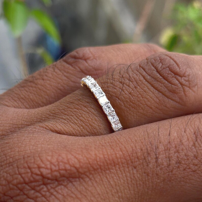 May include: A silver-toned band ring featuring a continuous row of square-cut clear stones set in a channel setting. The ring is worn on a finger, showcasing the metallic finish and the uniform arrangement of the faceted stones along the band.