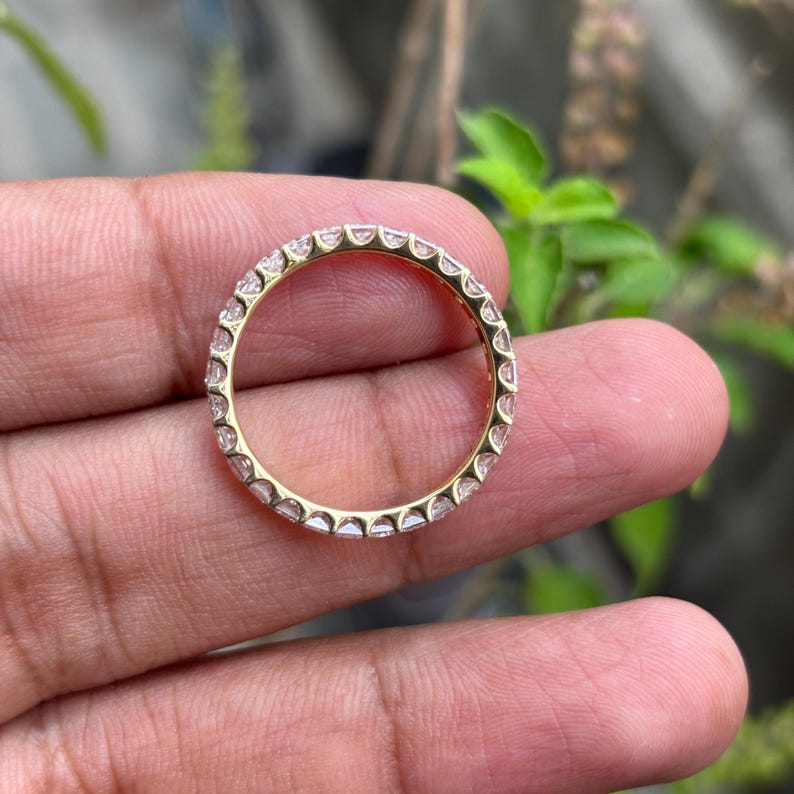 May include: A gold eternity band featuring a continuous row of small, clear, round-cut stones set in a shared-prong metal setting. The ring displays a uniform circular design with metallic prongs securing each individual stone around the entire band.