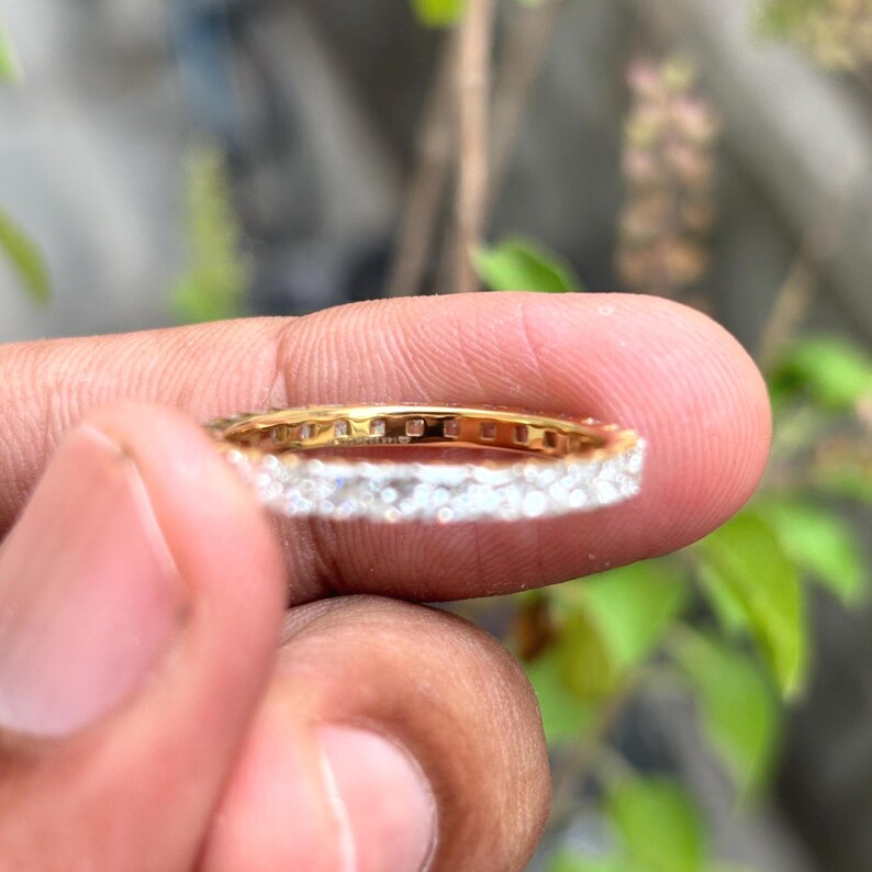 May include: A gold eternity band ring featuring a continuous row of small, clear, round-cut stones set along the outer circumference. The metal band has a polished yellow gold finish, and the stones are arranged in a shared-prong setting style.