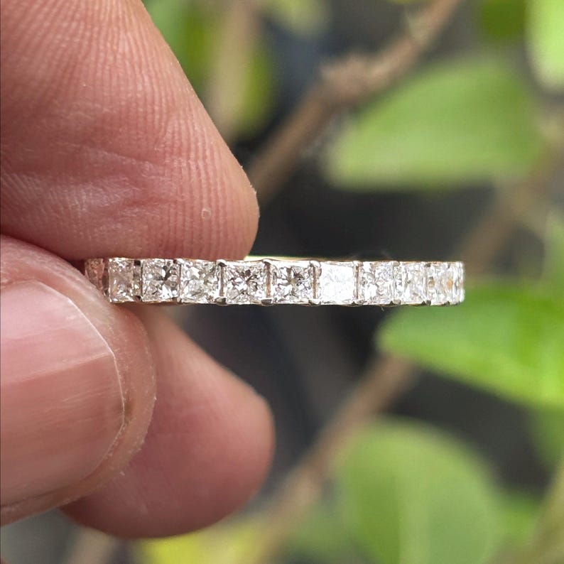 May include: A thin eternity band featuring a continuous row of small, square-cut clear stones set in a metallic frame. The ring displays a uniform arrangement of faceted gems that catch the light, creating a sparkling effect across the entire band.
