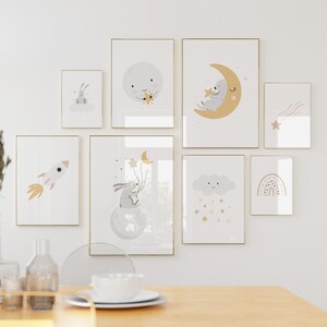 May include: A set of 8 framed prints featuring a whimsical nursery theme with illustrations of a bunny, moon, stars, clouds, a rocket, and a rainbow. The prints are in a minimalist style with a white background and gold frames.