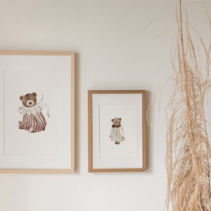 May include: Two framed illustrations of teddy bears wearing clothes. The larger illustration features a teddy bear wearing a pink and white striped dress with a white collar and bow. The smaller illustration features a teddy bear wearing a striped dress with a white collar.