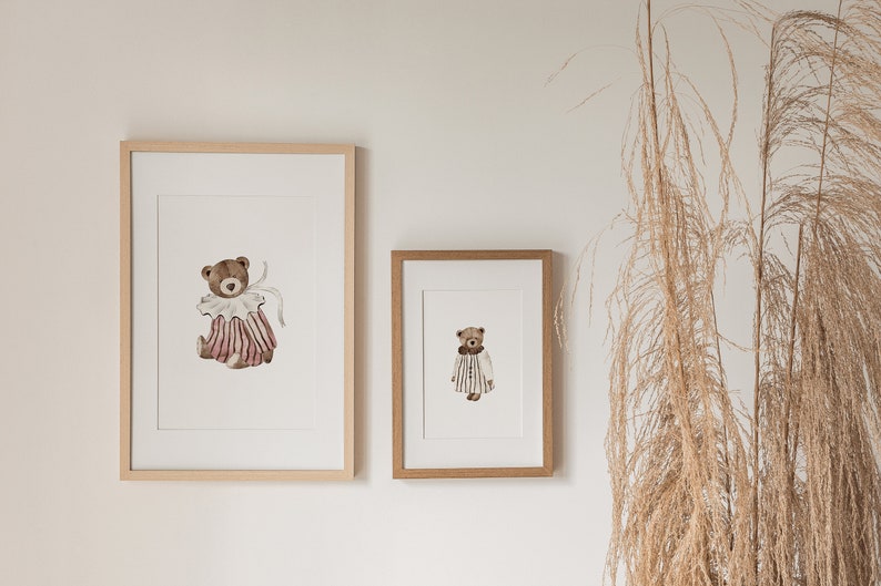 May include: Two framed illustrations of teddy bears wearing clothes. The larger illustration features a teddy bear wearing a pink and white striped dress with a white collar and bow. The smaller illustration features a teddy bear wearing a striped dress with a white collar.