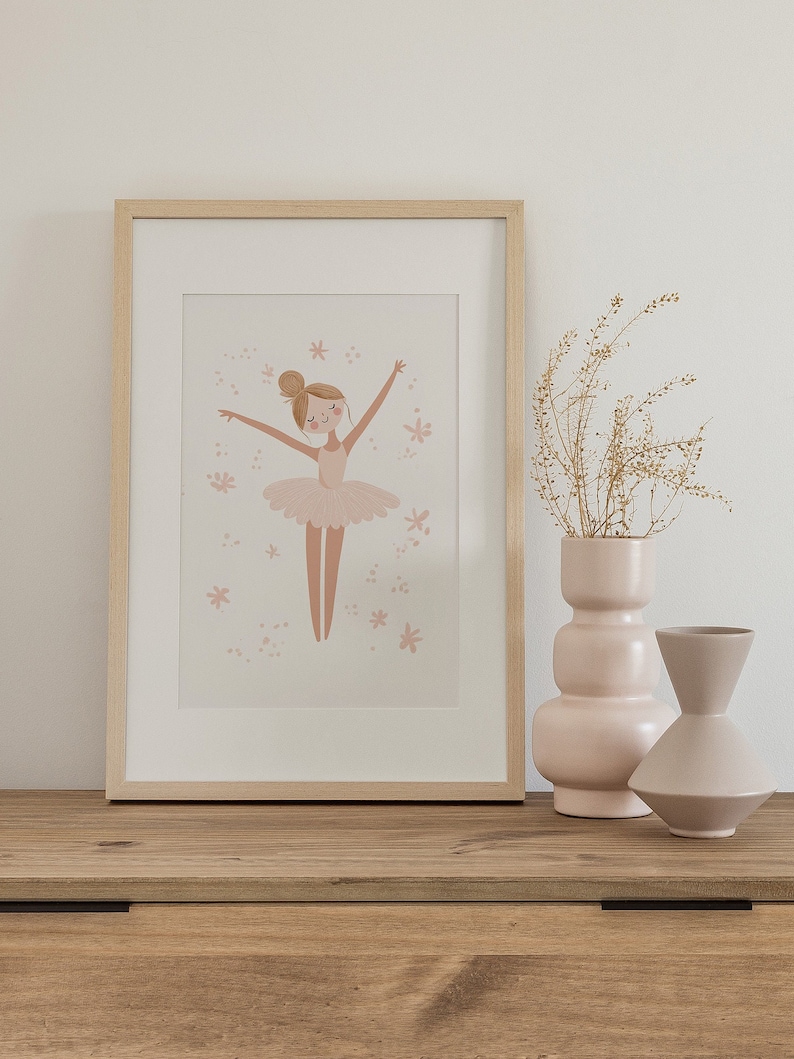 May include: A framed illustration of a ballerina in a pink tutu with a light pink background and white stars. The frame is light wood and is sitting on a wooden shelf with two pink ceramic vases.